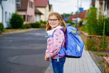 Happy little girl with glasses, wearing a pink sweater and a blue backpack walking to elementary school. She is smiling confidently, ready for school on a pleasant day.