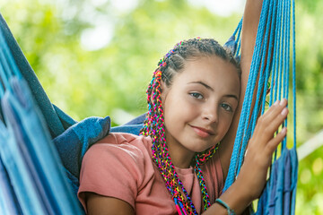 Beautiful young girl with colorful braids in her hair is posing on blue hammock.  Horizontally. 