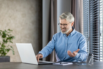 Fototapeta premium Senior businessman feeling frustrated due to malfunctioning laptop while sitting at modern office desk. Experiencing technical difficulties, visibly stressed. Surroundings include office plants