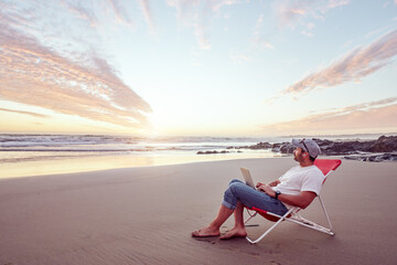 mature digital nomad sitting on the shore of the beach working with his laptop at beautiful sunset
