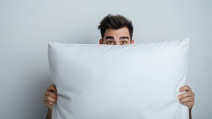 young man peeking from behind white pillow with wide eyes on gray background, concept of sleep, restlessness and calmness