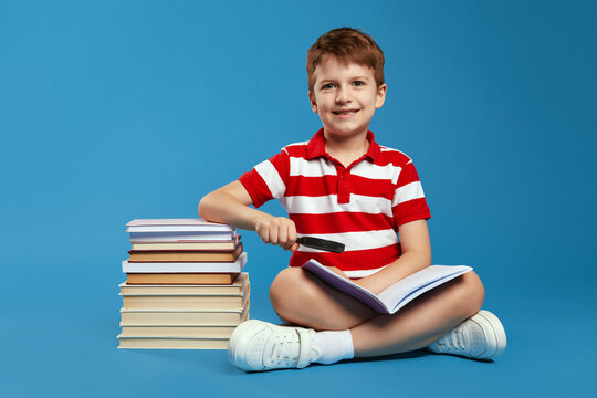 Curious child in red striped shirt smiling and using magnifier glass to read interesting book while sitting crossed legged on floor near bunch of books, isolated over blue background