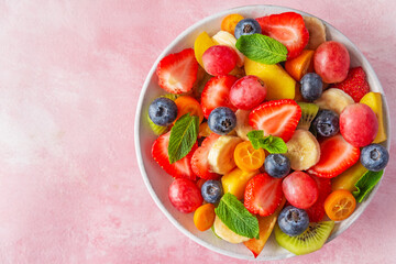 Healthy fresh fruit salad in a bowl on pink background. Top view with copy space. Summer healthy food for breakfast. Mixed fruits, berries and mint for diet lunch