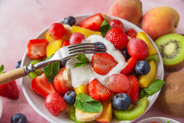 Summer fresh fruit salad with yogurt in a bowl with fork on pink background. Healthy food for breakfast. Mixed strawberries, grapes, banana, kiwi, blueberries, peach, kumquat and mint for diet lunch