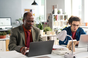 Portrait of African American senior intern working at laptop with young coworker in open office space copy space