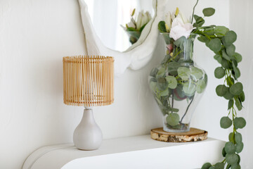 Scandinavian minimalistic interior design with table rattan lamp, eucalyptus branches in glass vase on shelf and mirror with textured white plaster frame on wall in light living room. Cozy home decor © Andriy Medvediuk