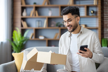 Young man excitedly opening a cardboard box while checking his smartphone at home