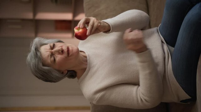 Vertical video: A woman who eats an apple while sitting on the couch has a toothache. She winces in pain as she takes a bite of the fruit, demonstrating the discomfort associated with toothache.
