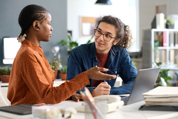 Portrait of smiling young man with long hair listening to female colleague discussing ideas during...