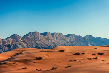 A vast, red sand desert stretches out in front of a range of rocky mountains against a clear blue sky. Photo view of desolation of the desert landscape. Orange sandy desert in Dubai