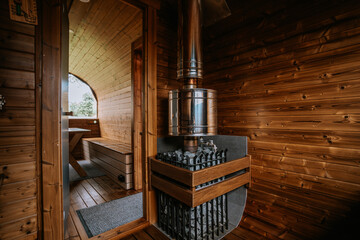 Cozy wooden sauna room with a shiny metal heater, benches, and a view of the outside through the open door. Curved wooden ceiling. Copy space.