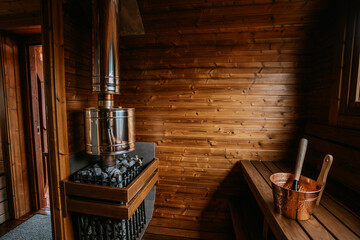 Wooden sauna interior with a shiny metal heater, a copper bucket and ladle on the bench, and wooden walls. Cozy and rustic atmosphere. Copy space...
