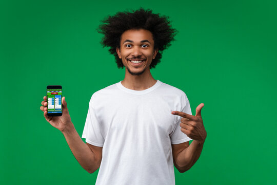 Young soccer fan man smiling and pointing with his index finger at smartphone with bookmaker's mobile application interface on its screen in hand
