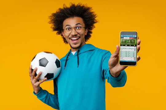 Portrait of happy smiling young man holding soccer ball in hand and reaching smartphone with bookmaker's application interface on its screen