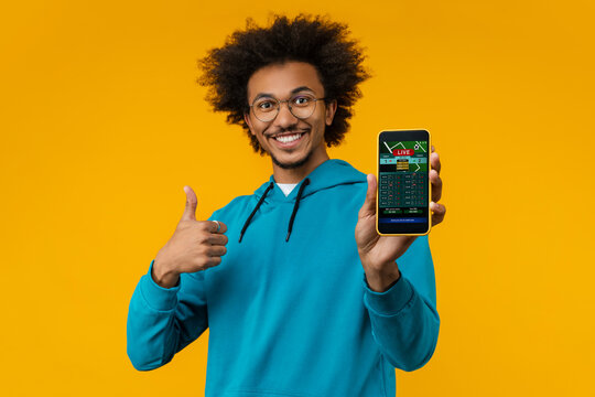 Studio shot of happy smiling young man showing thumbs up and reaching smartphone with bookmaker's application interface on its screen