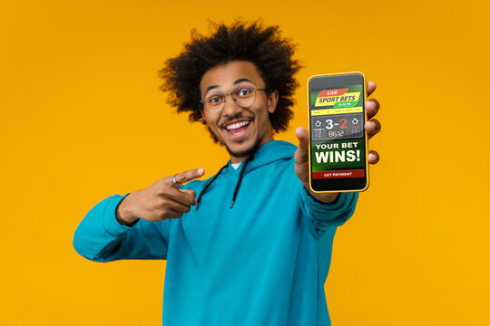 Portrait of happy smiling young African American man reaching smartphone with bookmaker's application interface on its screen