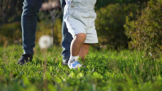 Father helps child take steps on grass. Child learning to walk outdoors. Happy bonding time with father. First steps in nature. Step, child, grass father moment. Happy family in the park. Happy family