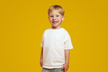 Cheerful little child in white t-shirt looking at camera with bright smile, standing isolated against yellow background. Children studio portrait.