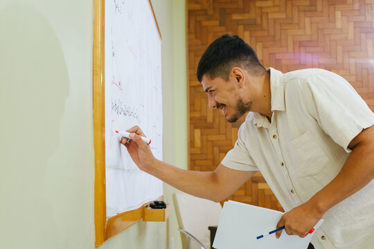 Latino teacher in classroom explaining concepts writing on blackboard.