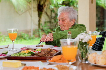 Elderly woman with white hair eating a meal sitting at a table outdoors.