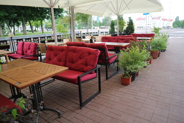 Cafe tables and chairs on the street. Under white umbrellas, there are wooden tables and red soft seats.