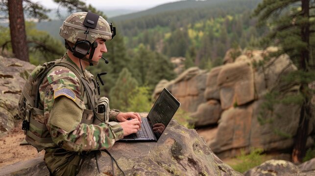 A soldier wearing a military uniform and helmet uses a laptop computer while sitting on a rock in a wooded area