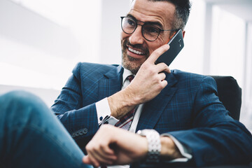 Successful entrepreneur middle aged laughing during phone conversation on smartphone sitting in office.Prosperous businessman in formal wear calling on cellphone.Cheerful proud ceo talking on cellular