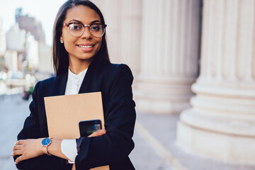 Half length portrait of cheerful dark skinned businesswoman in formal wear holding smartphone and...