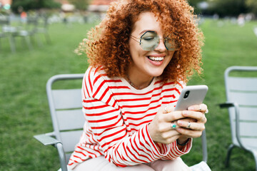 Outdoor portrait of hipster girl with red curly hair holding smartphone in hands sitting on chair in city park on lawn, chatting with her bestie or boyfriend, sending reels, sharing memes