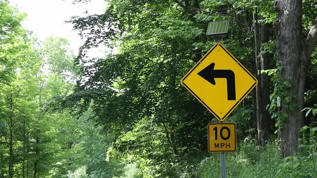 blinking traffic sign indicating a turn in the road with 10 mph (miles per hour speed limit signage) left arrow warning with lights on dangerous sharp curve on rural country street