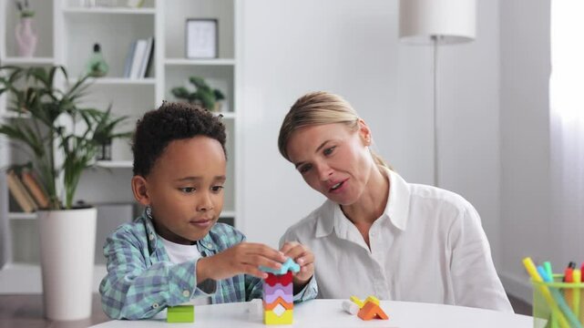 African American child curly little boy and child development specialist blonde woman playing with colorful wooden bricks sitting at table, making pyramid together. Exercises for children with autism.