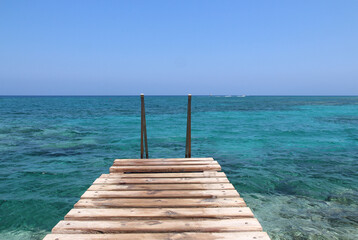 A wooden bridge with stairs, a pontoon bridge goes into the blue sea. A place for swimming, rest and relaxation. Turquoise clean transparent sea water of the Mediterranean Sea on a summer sunny day. 