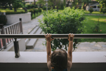 A child stands on a balcony, holding onto the railing and looking at the garden