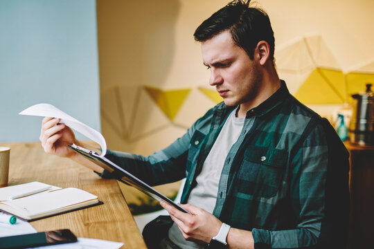 Pensive hipster guy reading report from planner and analyzing accountings for project upset with data, clever male coworker organizing information for creating architectural sketch concentrated