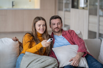 Obraz premium Contented man and woman watching TV together sitting on couch at home enjoying movies, shows, series. Pleased female holding remote control changing television channel. Family couple pastime, leisure,