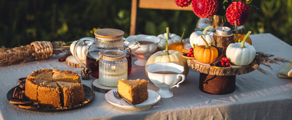 Tea setting with handmade pie. Beautiful view of garden wooden furniture with basket, soft blanket and burning candles. Outdoors fall family party, cozy autumn interior. Pumpkins, red dahlia banner