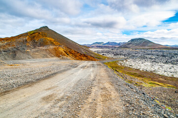 A dirt road winds through the stark and rugged landscape of solidified lava fields under a cloudy sky on Icelands Reykjanes Peninsula, showcasing striking contrasts in the terrains color and texture.