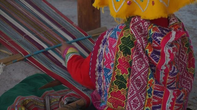 A woman is weaving a colorful blanket. The blanket is made of different colored strips of fabric. The woman is wearing a yellow scarf and a yellow hat