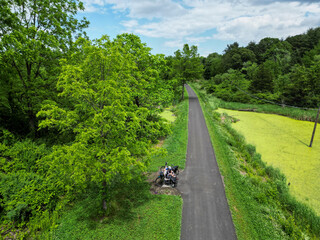 couple of cyclists sitting on bench at bike trail next to pond swamp (two people biking resting hudson valley path) paved bicycle road rural area