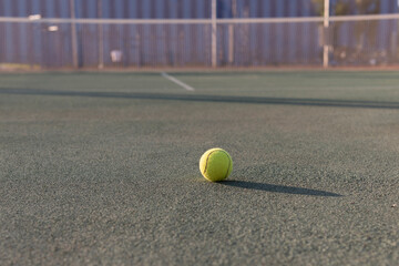 yellow tennis ball sits on a green tennis court, with a blurred background of a tennis net and trees