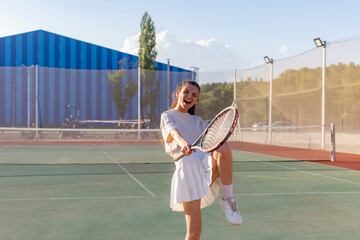 woman in a white tennis outfit celebrates a win with her racket raised high on a green tennis court