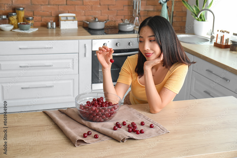 Poster Happy beautiful young Asian woman with bowl of ripe cherries in kitchen