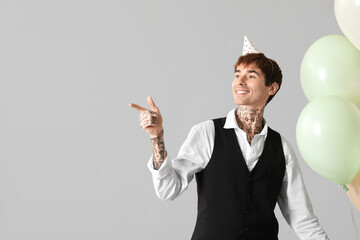 Young man in party hat with air balloons celebrating Birthday and pointing at something on grey background
