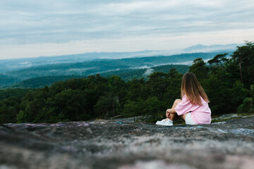 A young girl in white shorts, a pink T-shirt and white sneakers sits on a rock and looks at the mountains on a summer day. Tourist in the mountains. Dawn in the mountains. Hiker looks at sunrise