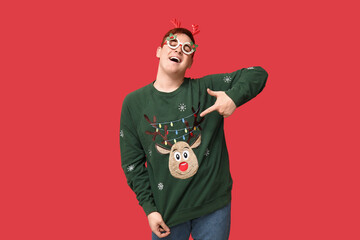 Handsome young man Christmas novelty glasses and reindeer headband pointing at his warm sweater on red background