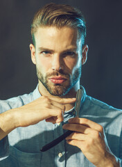 Barbershop. Professional hairdresser with straight razor. Serious barber shows sharp blade of dangerous razor. Handsome bearded man in denim shirt with classic old-fashioned sharp straight razor.