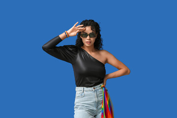 Young African-American woman with rainbow flag and stylish sunglasses on blue background. LGBT...