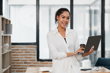 Smiling young woman in a white shirt holding a laptop and coffee cup in a modern office with large windows and brick wall.