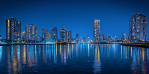 Naklejka premium A vibrant city skyline at night, with high-rise buildings reflecting off the calm water. The clear blue sky and illuminated windows create a lively yet serene urban scene, nightlife