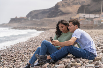 Fototapeta premium Couple sitting on the beach using phone